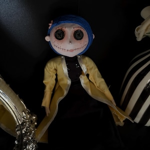 K&ouml;nnte beinhalten: Puppe mit blauen Haaren, Knopfaugen und einem gen&auml;hten L&auml;cheln, die einen gelben Mantel &uuml;ber einem schwarzen Kleid tr&auml;gt. Die Puppe hat eine rosa Blume im Haar und ist vor einem dunklen Hintergrund platziert.