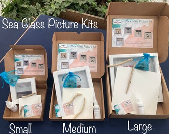 Könnte beinhalten: Drei braune Kartons in verschiedenen Größen, beschriftet mit "Klein", "Mittel" und "Groß". Jede Schachtel enthält ein Meerglass-Bilder-Set mit einem weißen Rahmen, einem Beutel mit Meerglas, einem Stück Treibholz und Kleber. Die Schachteln stehen auf einer blauen Oberfläche.