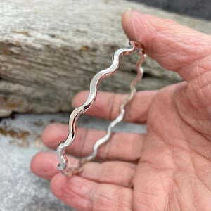 May include: A silver bangle with a distinctive wavy pattern. The bracelet is held in a hand, highlighting its polished finish. The design of the bangle is a series of gentle curves, giving it a contemporary look. The bracelet is a piece of jewellery.