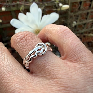 May include: A silver ring with a knot design. The ring is worn on a finger, with a blurred background of white flowers and a wooden fence. The ring's design features a central knot and a wavy band.