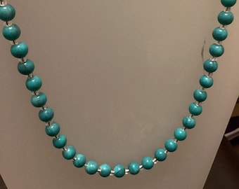 Puede incluir: Un collar de cuentas turquesa con detalles plateados. El collar está en un expositor blanco. El expositor tiene una etiqueta que dice "Colourful Creations Joyería artesanal".