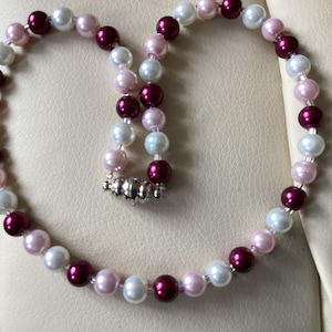 May include: A necklace composed of white, pale pink, and deep burgundy beads. The beads are strung together, with a silver-coloured clasp. The necklace is a classic design, with the beads evenly spaced and round in shape.