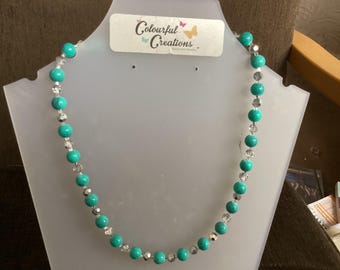 Puede incluir: Un collar de cuentas turquesa y plata en un expositor blanco. El collar presenta cuentas turquesas y transparentes alternadas. El expositor tiene una etiqueta que dice "Colourful Creations".