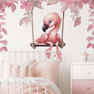 May include: Pink flamingo wall decal with watercolor leaves and flowers, sitting on a swing. The decal is on a white wall with a white bed and pink pillows in the foreground.