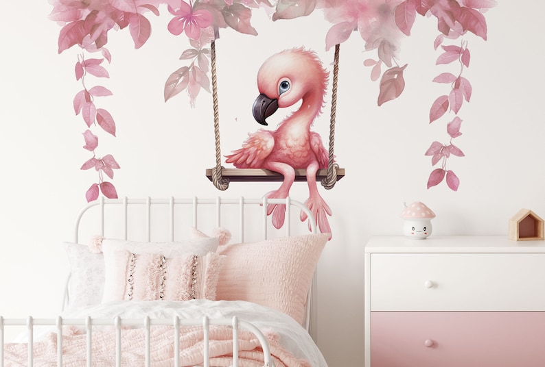 May include: Pink flamingo wall decal with watercolor leaves and flowers, sitting on a swing. The decal is on a white wall with a white bed and pink pillows in the foreground.