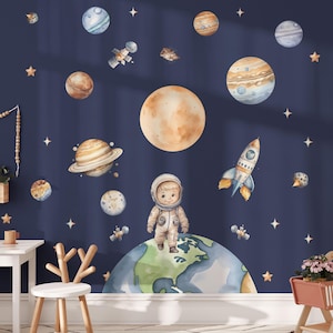 Può includere: Un muro blu con un adesivo murale a tema spaziale che presenta un astronauta dei cartoni animati in piedi su un pianeta, un razzo, pianeti, stelle e un'astronave.