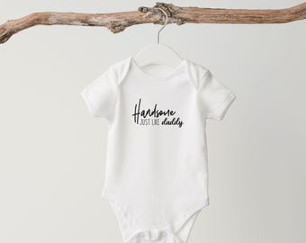 May include: White baby bodysuit with the words "Handsome just like daddy" in black script. The bodysuit hangs from a white hanger, suspended from a brown branch.