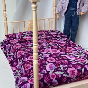 Puede incluir: Una cama con dosel en miniatura con una colcha y una almohada estampadas con flores. La ropa de cama presenta un fondo morado oscuro con diseños de flores rosas, moradas y azules. El marco de la cama es de color beige claro.