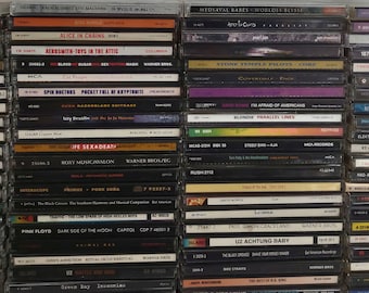 Może przedstawiać: Widok z bliska półki z kolekcją płyt kompaktowych (CD) ułożonych pionowo. Płyty CD są głównie czarne z białym tekstem i logo. Niektóre płyty CD mają kolorowe grafiki. Płyty CD są uporządkowane według gatunku, z rockiem, metalem i muzyką alternatywną jako najbardziej dominującymi gatunkami. Płyty CD są wszystkie w ochronnych etui.