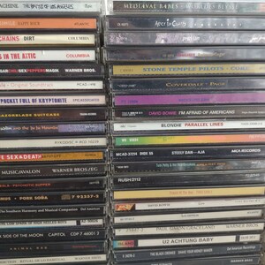 Może przedstawiać: Widok z bliska stosu kaset magnetofonowych z r&oacute;żnymi artystami i tytułami album&oacute;w. Kasetki są ułożone pionowo, grzbietami do widza. Niekt&oacute;rzy z przedstawionych artyst&oacute;w to Alice in Chains, Stone Temple Pilots, David Bowie i Blondie. Kasetki są połączeniem czarnych, białych i kolorowych pudełek.
