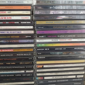 May include: A close-up view of a stack of cassette tapes, featuring various artists and album titles. The tapes are arranged vertically, with the spines facing the viewer. Some of the artists featured include Alice in Chains, Stone Temple Pilots, David Bowie, and Blondie. The tapes are a mix of black, white, and colored cases.
