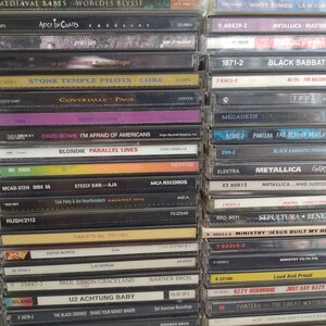 May include: A close-up view of a stack of compact discs (CDs) with various album covers. The CDs are arranged vertically, with the spines facing the viewer. The album covers feature a variety of artists and bands, including Metallica, Black Sabbath, and Rush. The CDs are all in black cases with silver lettering.