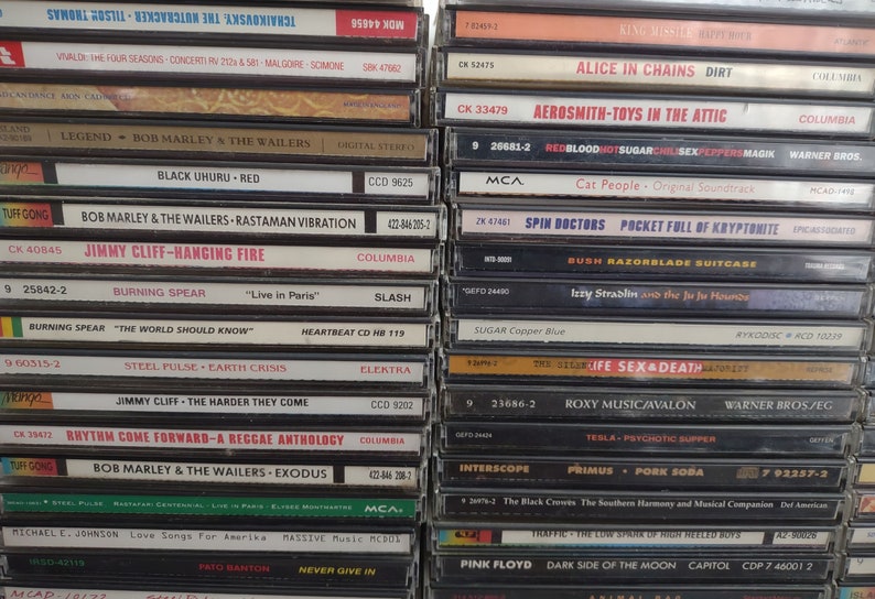 May include: A close-up view of a stack of compact discs (CDs) with various album covers. The CDs are arranged vertically, with the spines facing the viewer. The album covers feature a variety of artists and genres, including rock, pop, and reggae. Some of the album titles visible include "Alice in Chains", "Aerosmith", "Bob Marley", "Burning Spear", "Jimmy Cliff", "Roxy Music", "Pink Floyd", and "Spin Doctors".