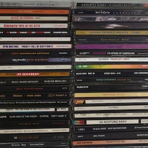 May include: A close-up view of a shelf with a collection of compact discs (CDs) stacked vertically. The CDs are mostly black with silver or white text and logos. Some of the CDs have colorful artwork. The CDs are organized by genre, with rock, metal, and alternative music being the most prominent. The CDs are a mix of new and old releases, with some of the CDs being from the 1990s and 2000s.