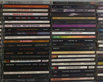 May include: A close-up view of a shelf with a collection of compact discs (CDs) stacked vertically. The CDs are mostly black with silver or white text and logos. Some of the CDs have colorful artwork. The CDs are organized by genre, with rock, metal, and alternative music being the most prominent. The CDs are a mix of new and old releases, with some of the CDs being from the 1990s and 2000s.