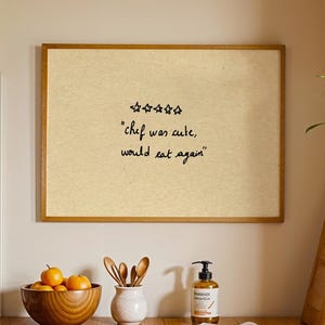 May include: A rectangular wall art piece in a light wood frame featuring a beige background with five hand-drawn stars and the black text "chef was cute, would eat again" centered on the print, displayed in a kitchen setting with wooden counters.