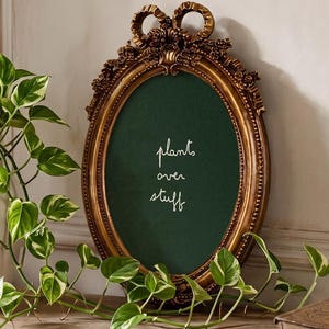 May include: An oval-shaped gold picture frame with ornate detailing and a bow-shaped top, containing a dark green insert with the white text "plants over stuff" written in a cursive script, positioned next to a white vase with a trailing plant.