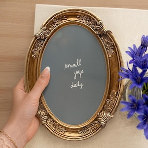 May include: An oval-shaped picture frame with an ornate, antique gold-colored finish and decorative floral relief details. The center features a gray background with the white handwritten text "small joys daily" centered in the middle of the frame.