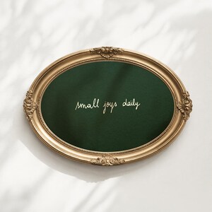 May include: An oval-shaped wall decor piece featuring a dark green velvet-like center enclosed in an ornate gold-colored frame with intricate leaf and scroll detailing. The text "small joys daily" is written in white cursive script in the center.