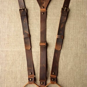 Può includere: Bretelle in pelle marrone con un design a testa di cervo sul davanti. Le bretelle hanno fibbie regolabili e passanti in pelle in basso.
