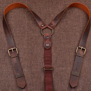 Puede incluir: Primer plano de tirantes de cuero marrón con hebillas de color latón y un anillo de conexión. Los tirantes tienen un diseño clásico con correas ajustables y una apariencia rica y texturizada. El cuero parece desgastado.