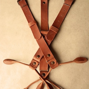 May include: Brown leather braces with adjustable straps and metal buckles. The braces have a Y-shaped back and feature a decorative ring at the top.