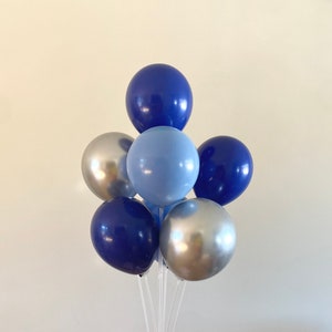 May include: A cluster of seven balloons in shades of blue and silver. The balloons are arranged in a bouquet, with a light blue balloon in the center, surrounded by navy blue and silver balloons. The balloons are attached to clear plastic sticks.