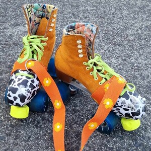 May include: A pair of orange roller skates with black and white cow print wheels and a bright orange strap with yellow daisy print. The skates have green laces and a colorful patterned lining.