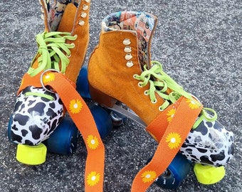Puede incluir: Un par de patines de ruedas naranjas con ruedas de estampado de vaca en blanco y negro y una correa naranja brillante con estampado de margaritas amarillas. Los patines tienen cordones verdes y un forro estampado de colores.
