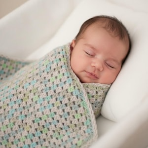May include: A sleeping baby wrapped in a handmade crochet blanket. The blanket features a granny square pattern in shades of gray, blue, and green. The baby is resting on a white pillow in a white bassinet.