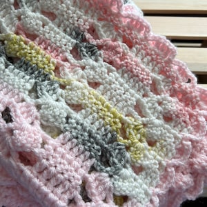 May include: A crocheted baby blanket in pink, white, gray, and yellow. The blanket has a scalloped edge and a delicate, lacy pattern.