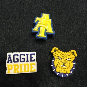 Könnte beinhalten: Drei Schuhanhänger aus Gummi mit College-Motiven. Ein Anhänger zeigt die Buchstaben "A" und "T" in Gelb auf blauem Hintergrund. Ein anderer Schriftzug lautet "AGGIE PRIDE" in Weiß, Blau und Gelb. Der dritte ist ein gelber Bulldoggenkopf mit blauem Kragen.