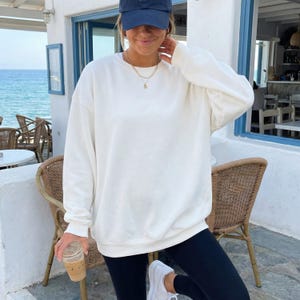 May include: An oversized white crewneck sweatshirt with long sleeves and ribbed cuffs, paired with black leggings, a navy blue baseball cap, and a delicate gold chain necklace. The sweatshirt features a simple, solid-colored design.