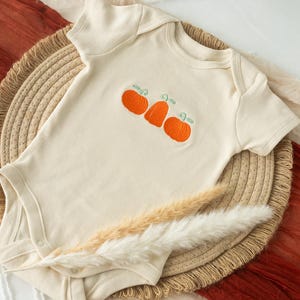 May include: A cream-colored baby onesie with a cute embroidered design of three orange pumpkins. The onesie is on a woven straw mat.