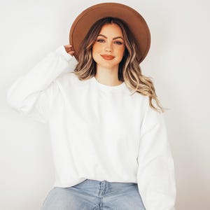 May include: A plain white long-sleeve crewneck sweatshirt paired with a wide-brimmed brown felt hat and light blue denim jeans. The sweatshirt features a simple, solid-colored design with long sleeves and a ribbed neckline, cuffs, and hem.