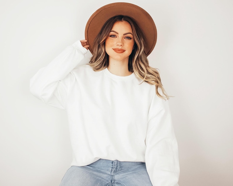 May include: A plain white long-sleeve crewneck sweatshirt paired with a wide-brimmed brown felt hat and light blue denim jeans. The sweatshirt features a simple, solid-colored design with long sleeves and a ribbed neckline, cuffs, and hem.