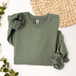 May include: A solid olive green crewneck sweatshirt with long sleeves and a ribbed collar. The garment features a Gildan brand label at the inner neckline. The fabric has a smooth, matte finish and is presented in a folded, relaxed arrangement.