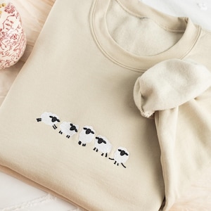 May include: A beige sweatshirt with a crew neck and long sleeves. The sweatshirt features an embroidered design of five sheep in a row. The sheep are white with black faces and legs. The sweatshirt is folded on a light-colored surface.