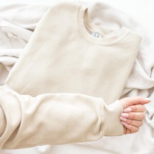 May include: A solid beige crewneck sweatshirt featuring long sleeves with ribbed cuffs and a ribbed neckline. The garment is constructed from a smooth, plain fabric and is presented in a simple, unadorned style with a classic pullover design.