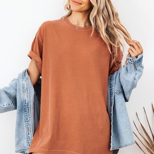 May include: A solid rust-colored crew neck t-shirt with short sleeves and a relaxed fit, worn layered underneath a light blue denim jacket. The t-shirt features a plain, unpatterned fabric design with a classic round neckline and rolled sleeves.
