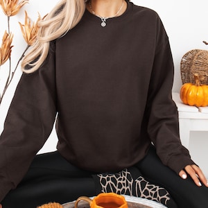 May include: A dark brown crewneck sweatshirt with long sleeves and a ribbed collar, cuffs, and hem. The garment features a solid, plain design with no visible patterns, logos, or text, providing a simple and uniform appearance throughout the fabric.