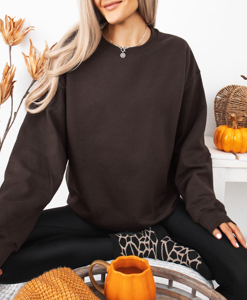 May include: A dark brown crewneck sweatshirt with long sleeves and a ribbed collar, cuffs, and hem. The garment features a solid, plain design with no visible patterns, logos, or text, providing a simple and uniform appearance throughout the fabric.