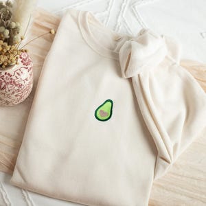 May include: Cream-colored sweatshirt with a small embroidered avocado design on the front. The avocado is green with a brown pit. The sweatshirt is folded, with the sleeves and hood visible. A small vase with dried flowers is in the background.