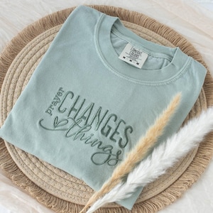 May include: A light green Comfort Colors crew neck t-shirt with the embroidered phrase "prayer changes things." The shirt is folded on a woven placemat, with dried decorative grasses. The shirt has a tag that reads "Comfort Colors".