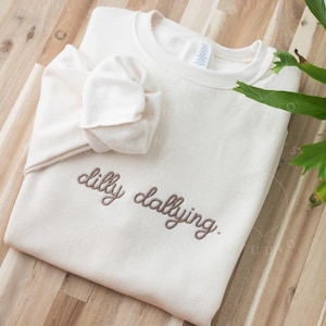 May include: Cream-colored sweatshirt with the embroidered phrase "dilly dallying" in a brown script. The folded sleeves add a casual touch. The sweatshirt is on a wooden surface.