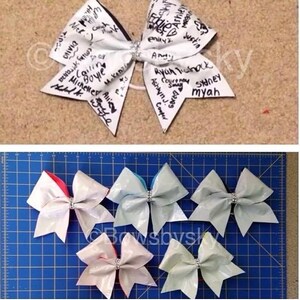 Op de afbeelding: Een witte cheer bow met zwarte tekst erop. De tekst bevat namen zoals "Nick", "Eddie", "Ryan Tishack" en "Sydney". Er zijn ook vijf kleinere cheer bows in verschillende kleuren op een blauw oppervlak.