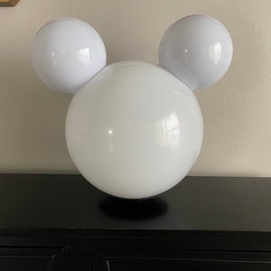 May include: White lamp with a round, white base and two round, white bulbs on either side, resembling Mickey Mouse ears.