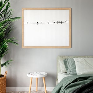 May include: A framed print featuring a minimalist design of several birds perched on a wire against a white background. The frame is light-colored wood, and the artwork is displayed on a gray wall. The scene evokes a sense of calm and nature.