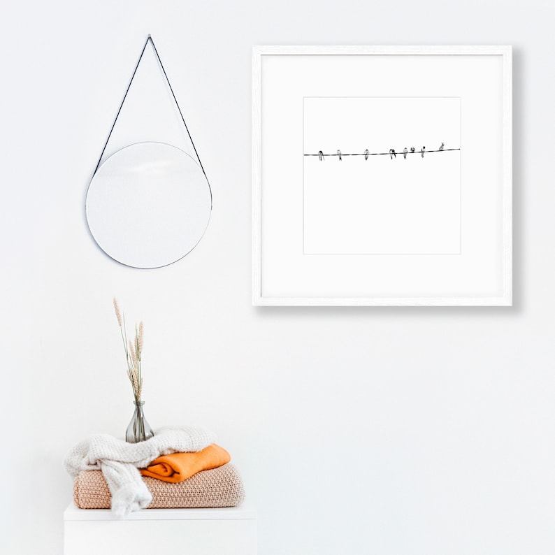 May include: A minimalist white framed print featuring a black line with several small bird silhouettes perched on it. A round mirror hangs on the wall, and a stack of folded blankets and a vase with dried grass are on a white surface.