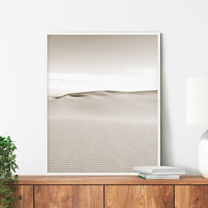 Könnte beinhalten: Ein gerahmter Druck mit einer minimalistischen Wüstenlandschaft. Das Kunstwerk zeigt eine Sanddüne unter einem gedämpften Himmel, mit einer neutralen Farbpalette aus Beige und Off-White. Der Druck ist in einem weißen Rahmen eingefasst, geeignet für die Inneneinrichtung.
